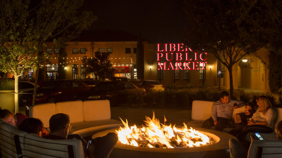 People relax around an outdoor fire pit at night in front of the glowing Liberty Public Market sign.