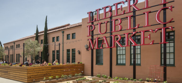 Large red Liberty Public Market sign outside a tan building with garden beds and an outdoor patio seating area