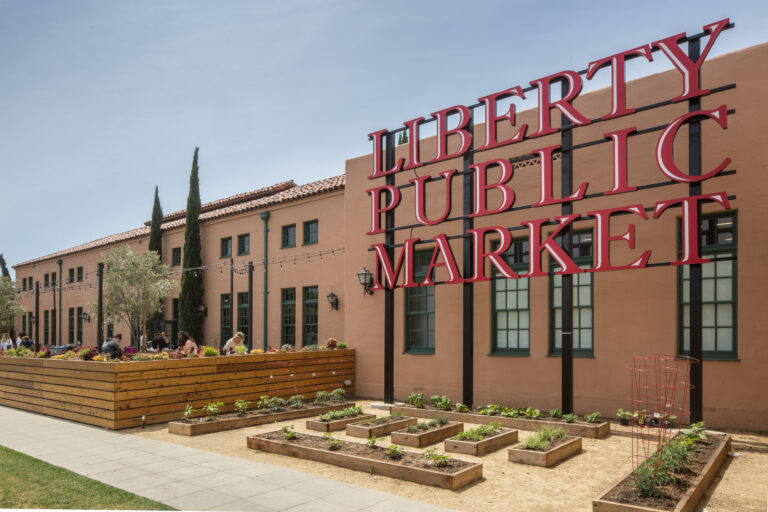 Large red Liberty Public Market sign outside a tan building with garden beds and an outdoor patio seating area