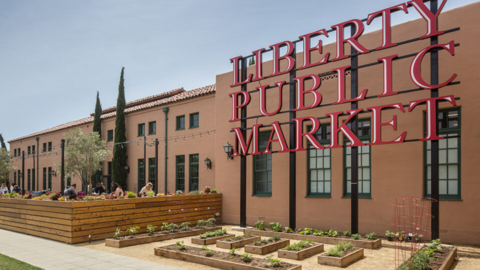 Large red Liberty Public Market sign outside a tan building with garden beds and an outdoor patio seating area