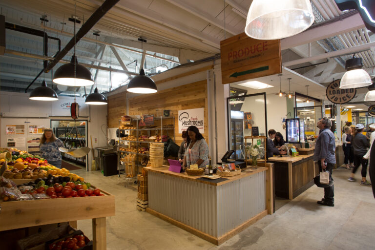 Indoor market with produce tables, a mushrooms stand, and shoppers under hanging lights.