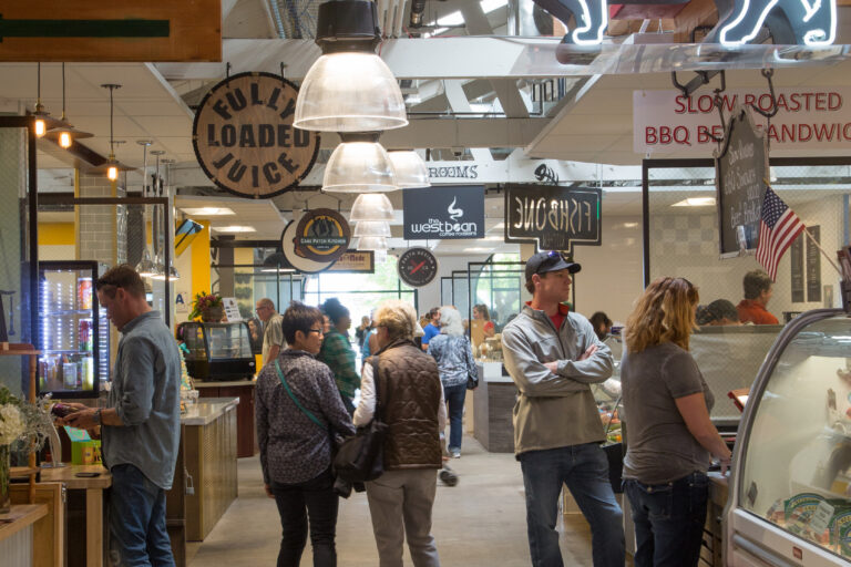 Shoppers in an indoor market aisle beneath hanging lights and a neon Liberty Meat Shop cow sign