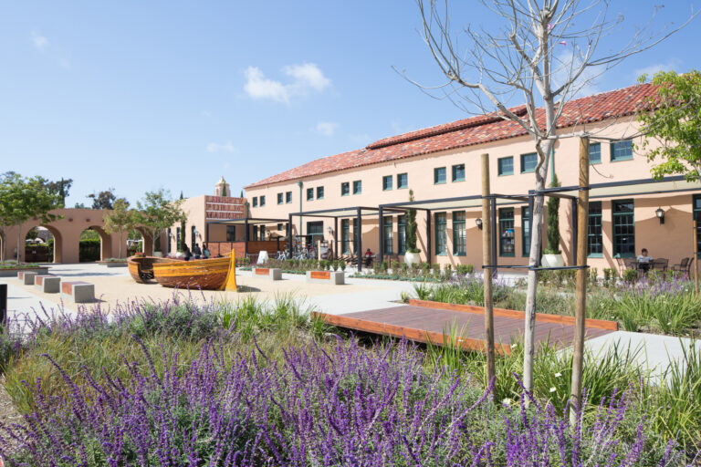 Liberty Public Market building with a tiled roof, landscaped courtyard, and wooden boat-shaped seating under a clear sky