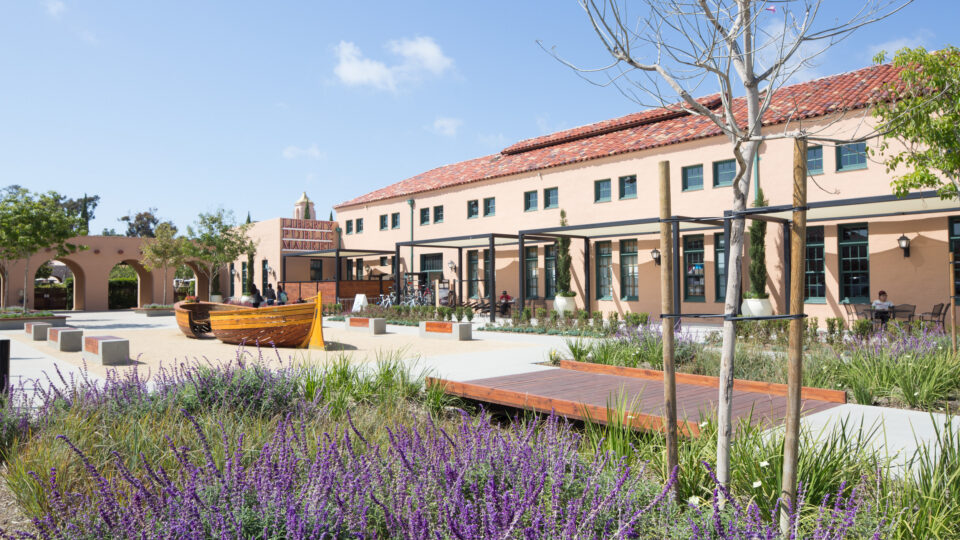 Liberty Public Market building with a tiled roof, landscaped courtyard, and wooden boat-shaped seating under a clear sky