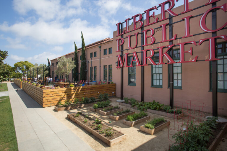 Outdoor seating and garden beds beside a building with a large red Liberty Public Market sign.