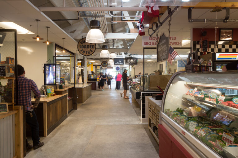 Indoor market aisle with food stalls, people walking, and a butcher’s meat display case on the right