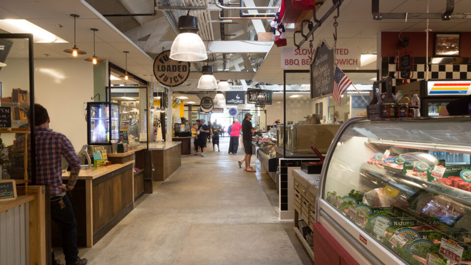 Indoor market aisle with food stalls, people walking, and a butcher’s meat display case on the right