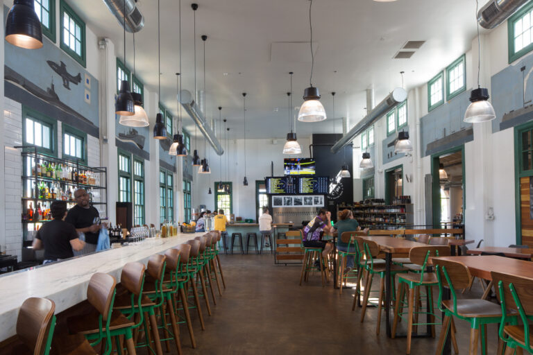 Spacious modern bar and restaurant interior with a long marble counter, green-framed windows, and nautical wall murals