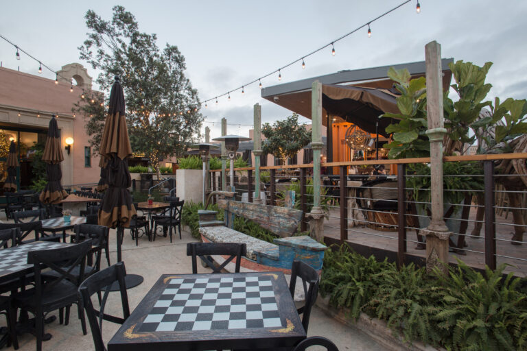 Outdoor restaurant patio at dusk with checkerboard tabletops, closed umbrellas, string lights, and lush plants