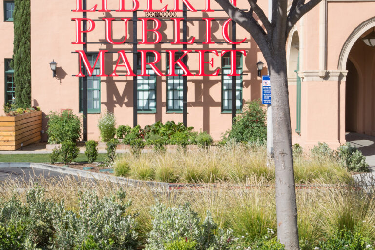 Red Liberty Public Market sign in front of a peach-colored building, partly obscured by a large tree and landscaping