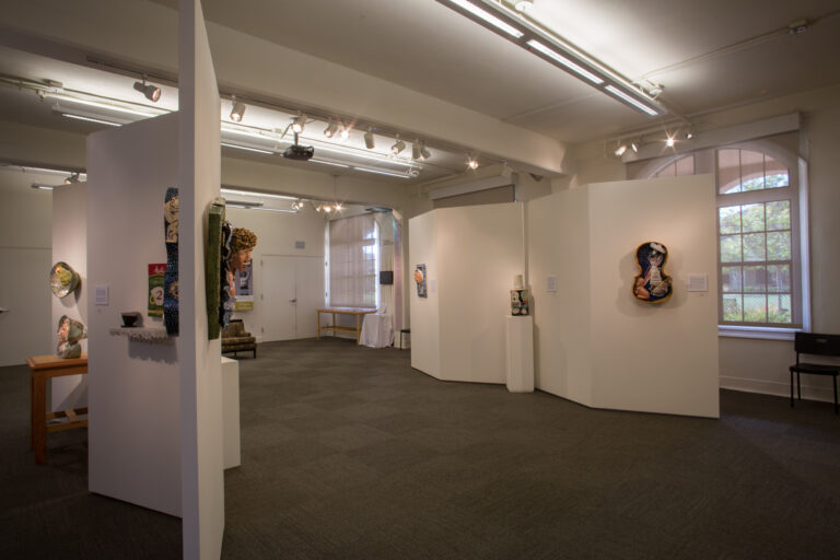 Wide view of an art gallery with white display walls, mixed-media artworks, spotlights, and tall arched windows.