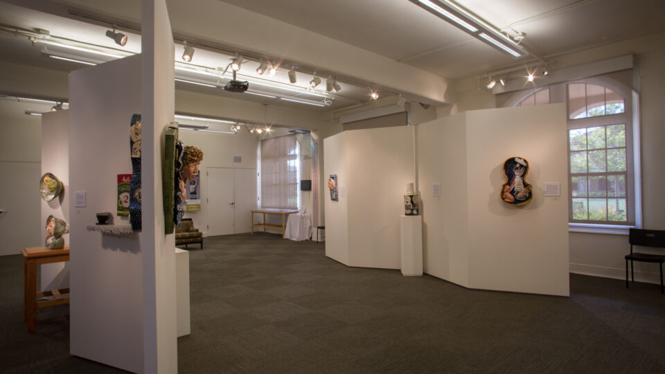 Wide view of an art gallery with white display walls, mixed-media artworks, spotlights, and tall arched windows.