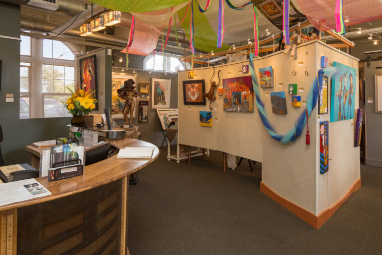 Art gallery interior with colorful paintings, sculptures, a curved front desk, and bright fabric decorations hanging overhead