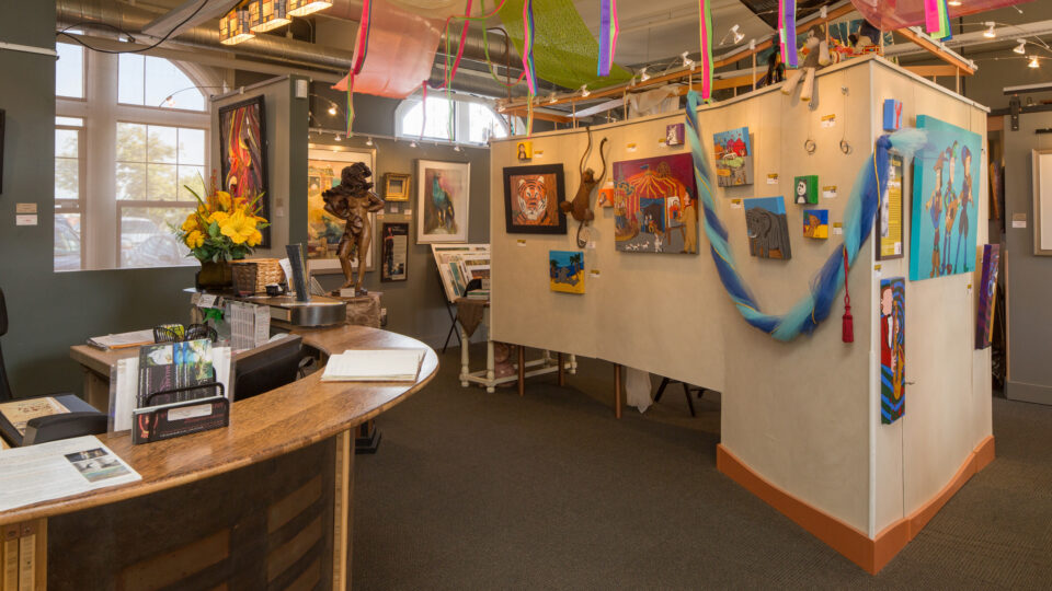 Art gallery interior with colorful paintings, sculptures, a curved front desk, and bright fabric decorations hanging overhead