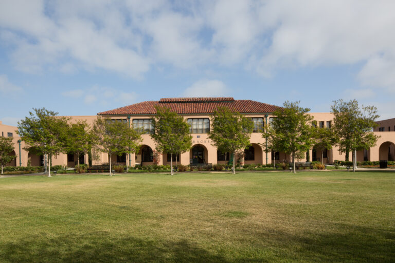 Spanish-style campus building with arched walkways, trees, and a large grassy lawn under a partly cloudy sky