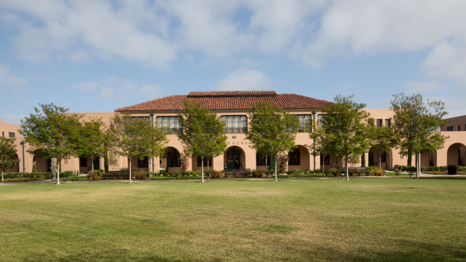 Spanish-style campus building with arched walkways, trees, and a large grassy lawn under a partly cloudy sky