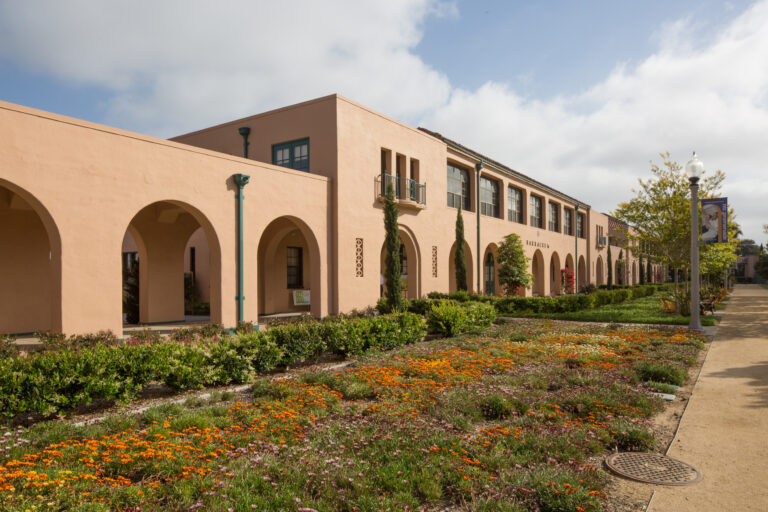 Spanish-style stucco building with arched walkways, landscaped flower beds, and a sidewalk under a partly cloudy sky
