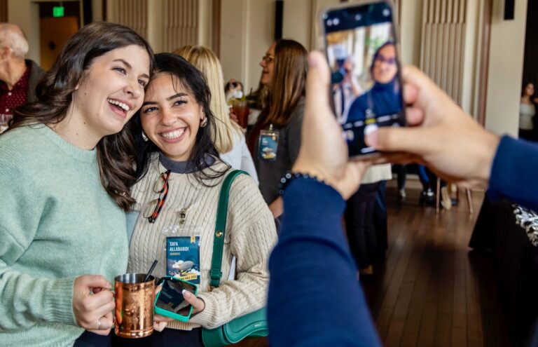 Two women smiling and posing for a smartphone photo at an indoor social event, one holding a copper mug.