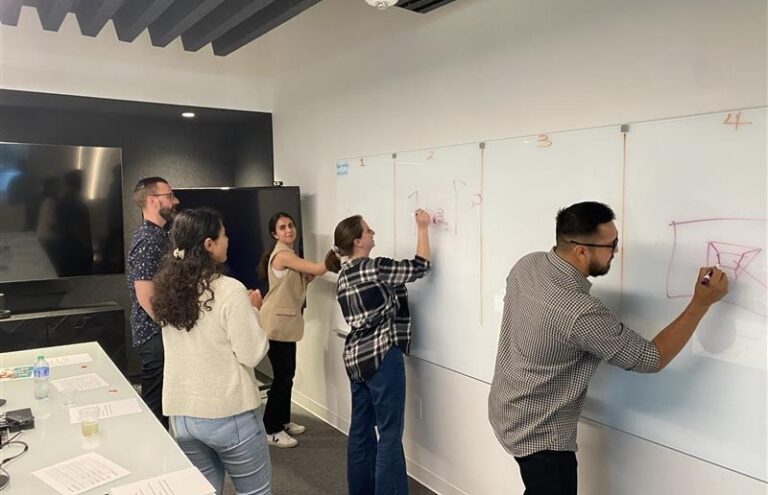 Five people in an office brainstorming at a whiteboard, drawing in separate sections during a workshop.