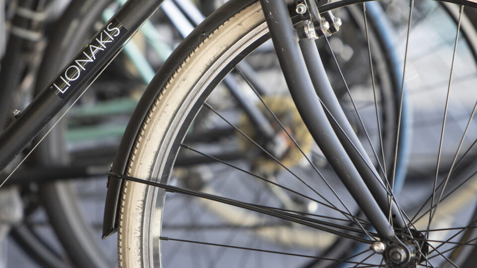 Close-up of a black bicycle’s front wheel and frame with the word LIONAKIS, with other bicycles blurred behind.