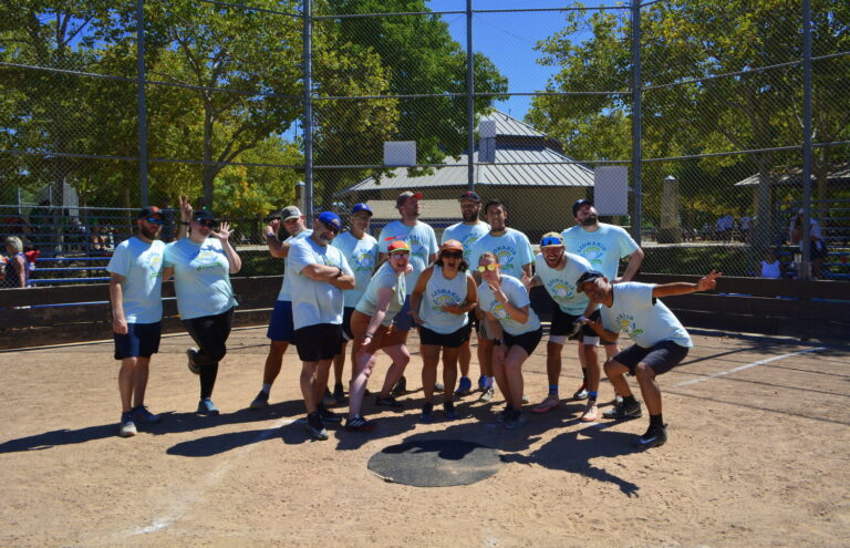 Group of adults in matching shirts posing playfully on a sunny baseball field in front of the backstop