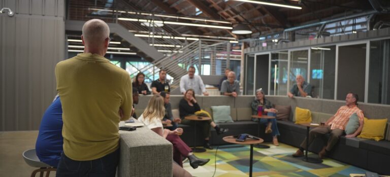 Group of coworkers gathered in a modern open office lounge, listening to a standing speaker during a casual meeting.