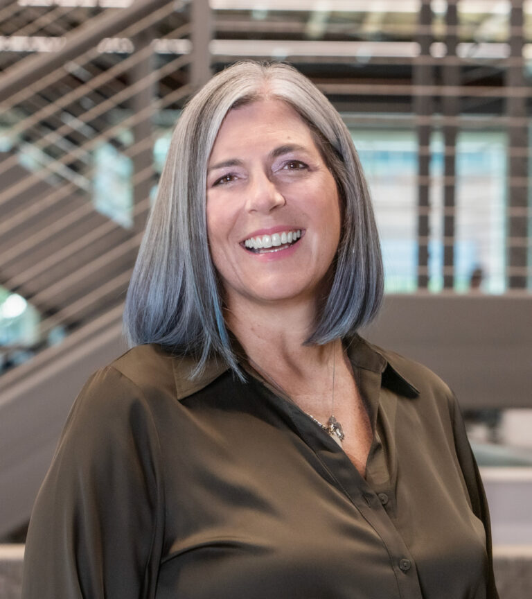 Smiling woman with shoulder-length gray hair in a dark blouse, standing indoors near a staircase.