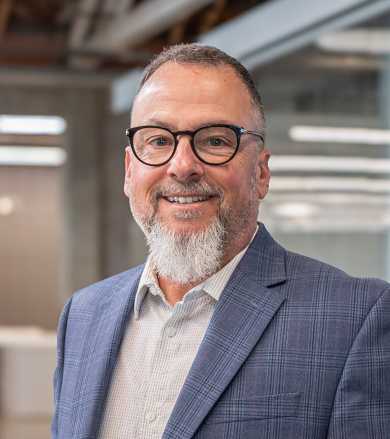 Smiling man with glasses and a gray beard wearing a blue plaid blazer in an office setting