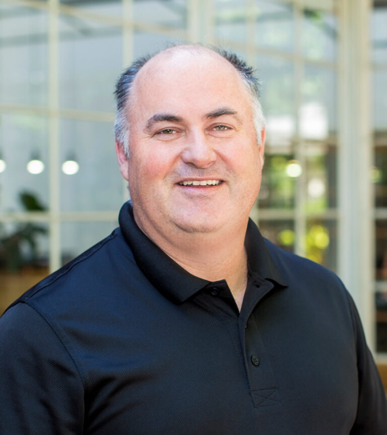 Smiling middle-aged man in a black polo shirt, posed indoors in front of large glass windows.