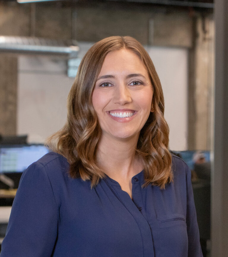 Smiling woman with shoulder-length brown hair in a blue blouse, standing in an office.