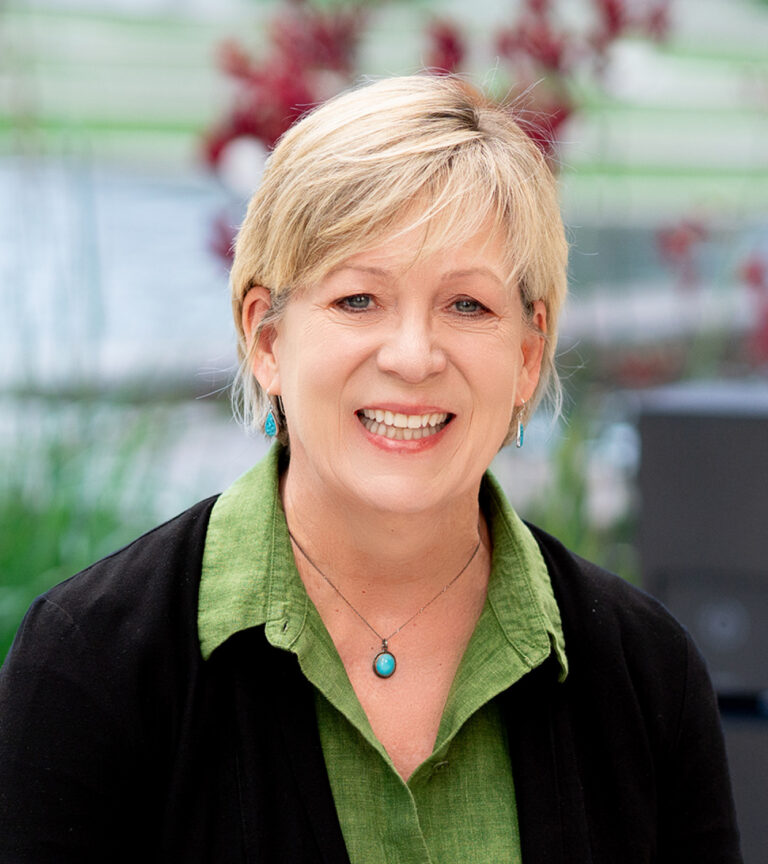 Smiling woman with short blond hair wearing a green shirt, black cardigan, and turquoise jewelry indoors