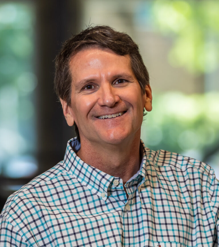 Smiling man in a blue and white checkered button-down shirt, with a softly blurred indoor background