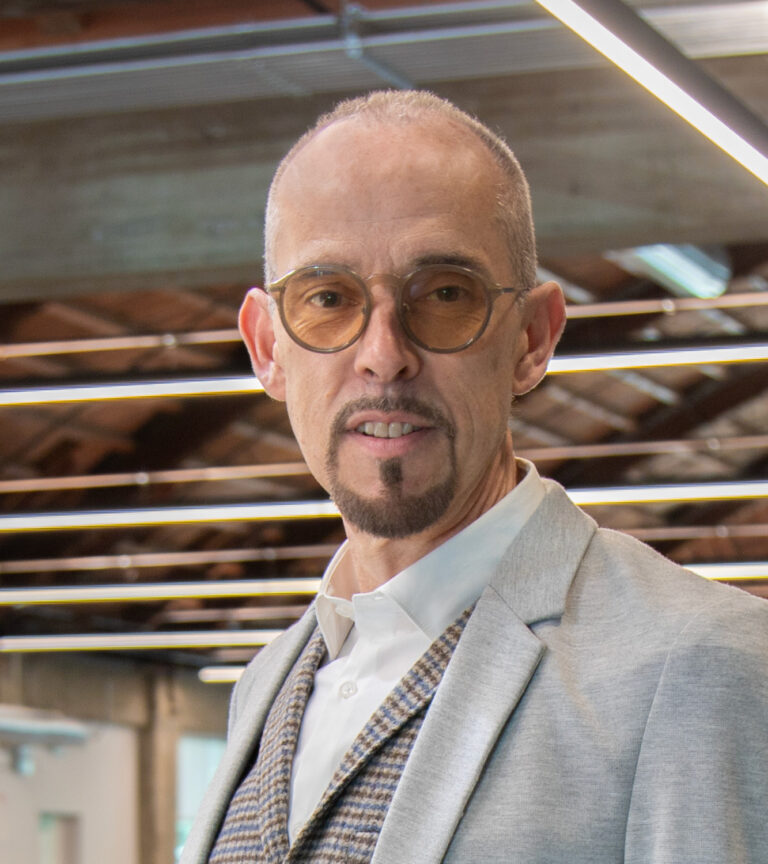 Portrait of a bald man with round glasses and a goatee, wearing a light gray blazer in a modern office.