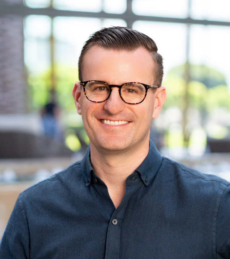 Smiling man with short dark hair and tortoiseshell glasses in a navy button-up shirt indoors