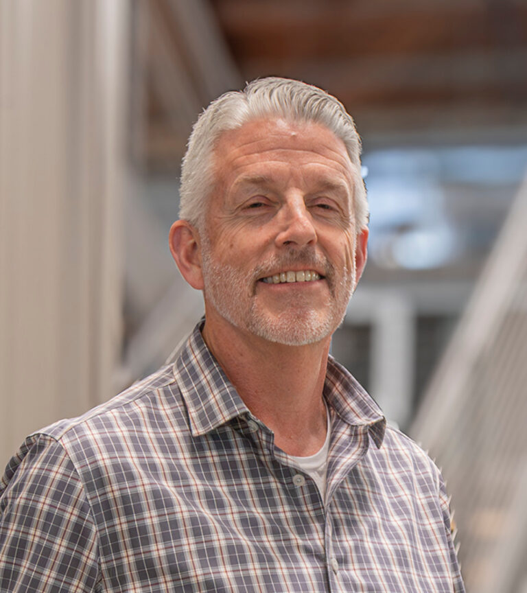 Smiling gray-haired man with a short beard wearing a plaid button-down shirt indoors.