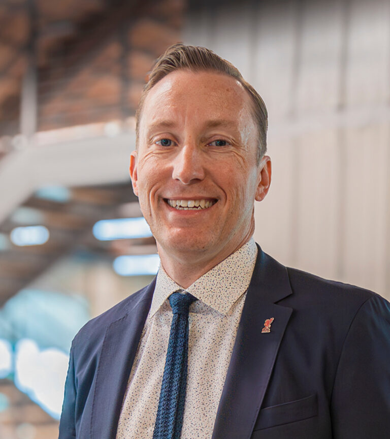 Smiling man in a navy suit and tie in a bright indoor setting with a blurred modern background