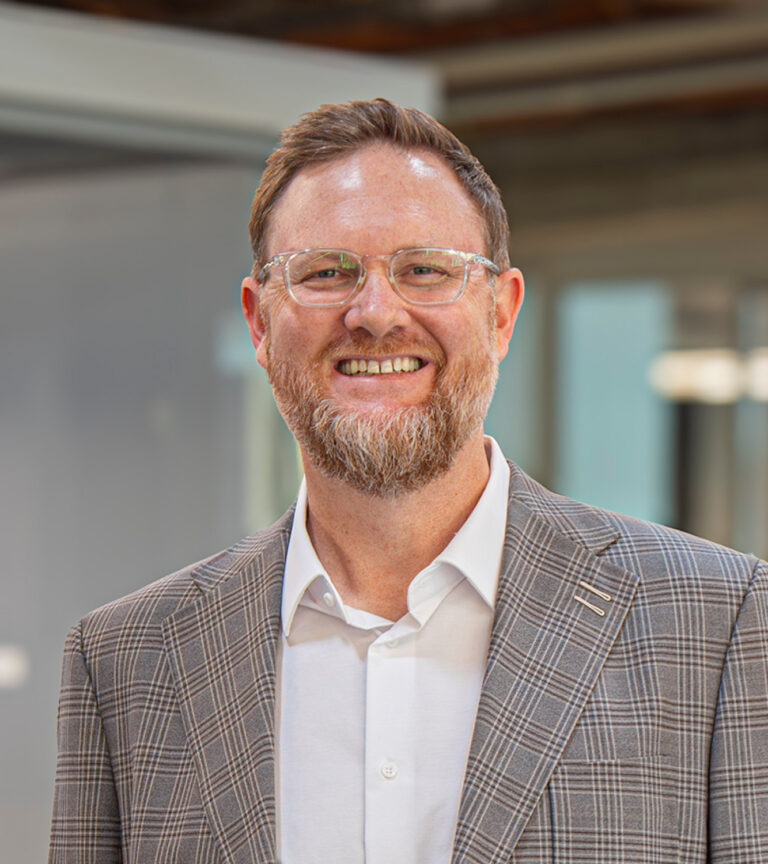 Smiling man with glasses and a beard wearing a gray plaid blazer and white shirt in an office setting