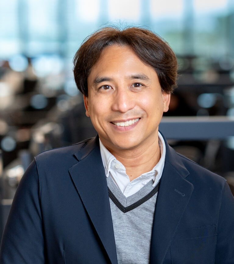 Smiling man in a navy blazer and gray sweater, posed indoors against a blurred office background