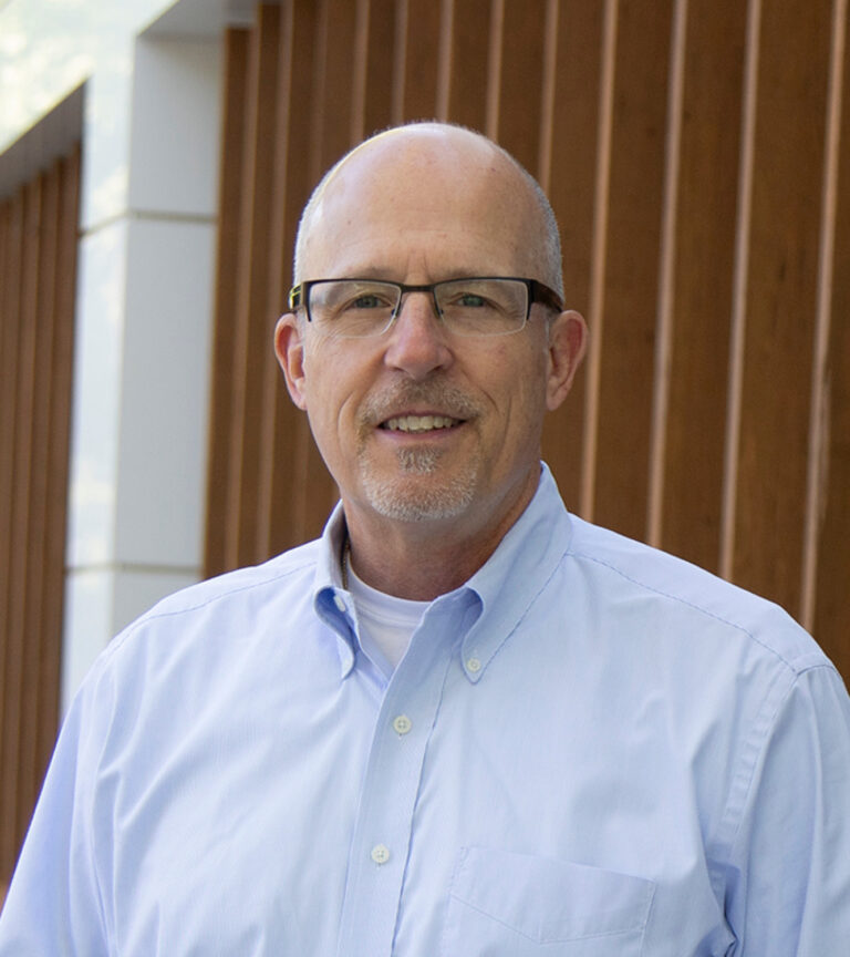 Smiling bald man with glasses and a goatee in a light blue button-down shirt, standing indoors against wood paneling