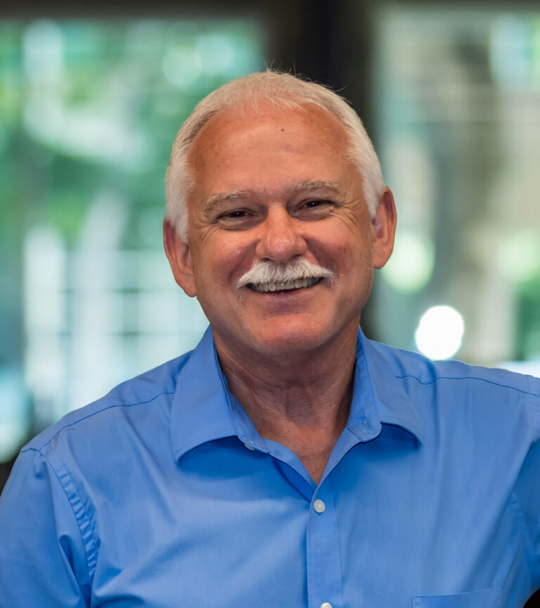 Smiling older man with white hair and a white mustache wearing a blue button-down shirt indoors
