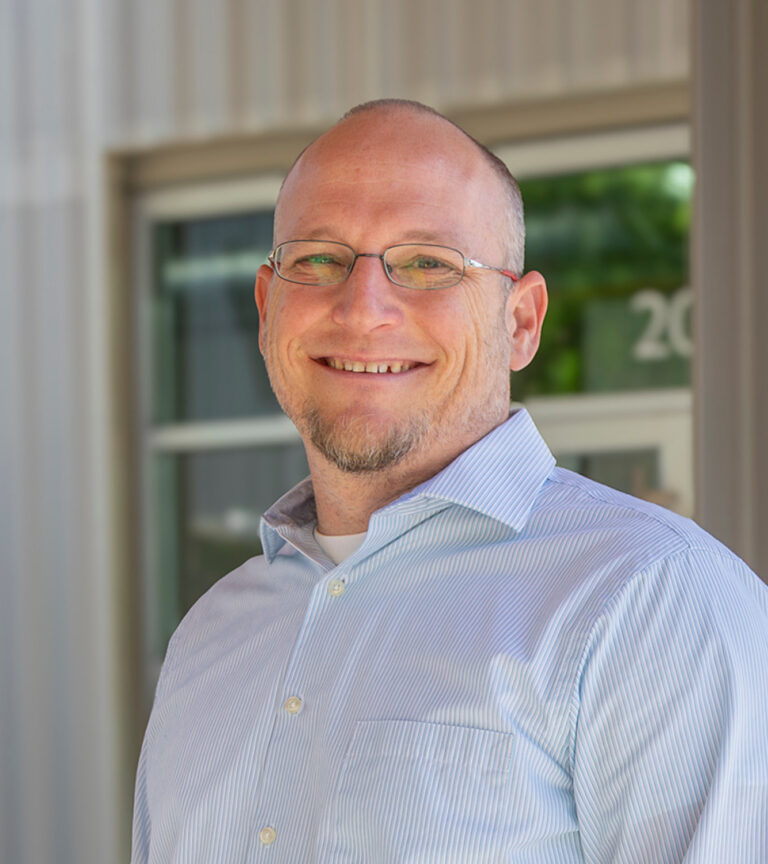 Smiling bald man with glasses and a short goatee wearing a light blue striped button-down shirt.