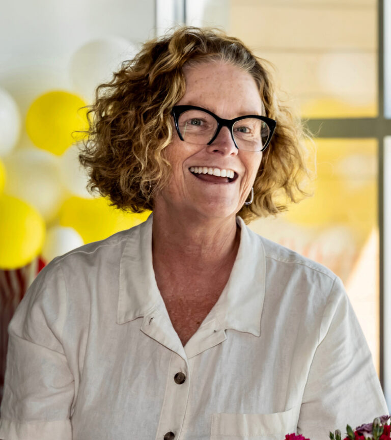 Smiling woman with curly hair and black glasses in a white blouse, with yellow and white balloons in the background
