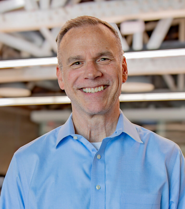 Smiling man in a light blue button-down shirt in an office with exposed beams and overhead lights