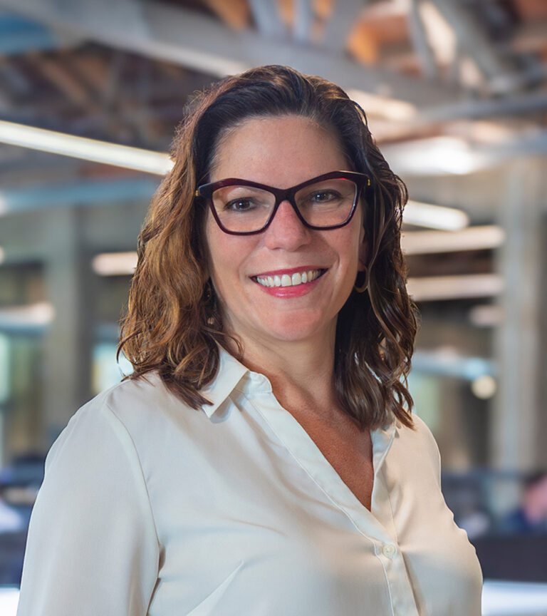 Smiling woman with wavy brown hair and dark glasses in a white blouse, standing in a modern office.