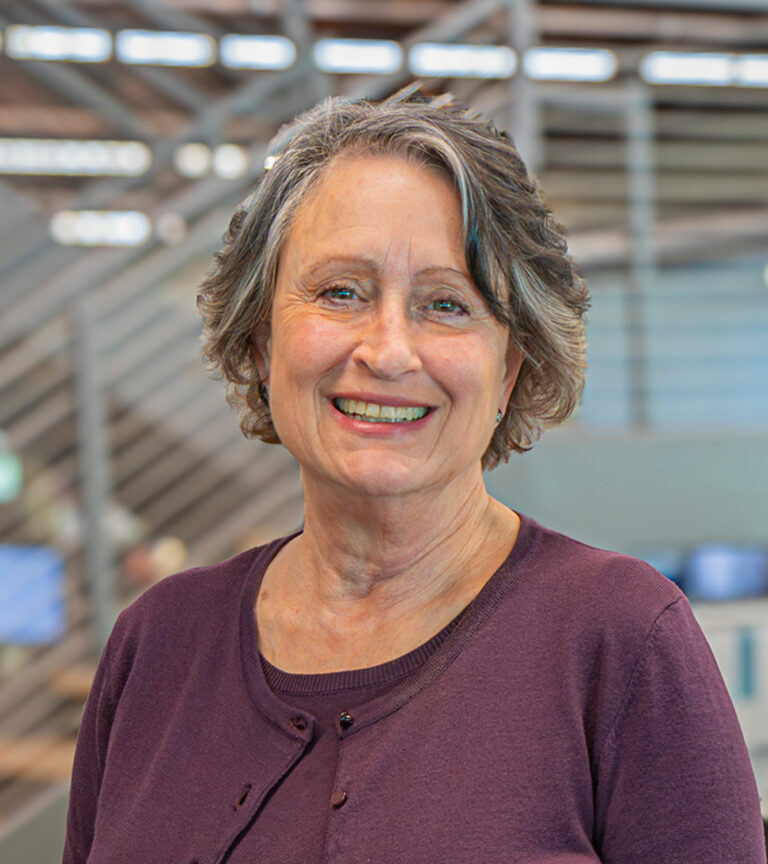 Smiling older woman with short gray hair in a purple cardigan, standing indoors against a blurred industrial background