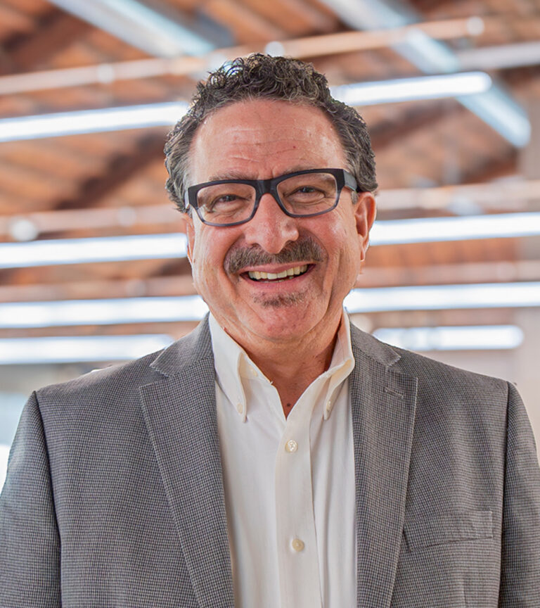 Smiling man with glasses, a mustache, and a gray blazer in a bright office setting