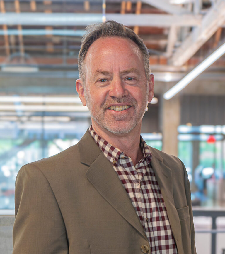 Smiling man in a tan blazer and checkered shirt standing in a modern office building.