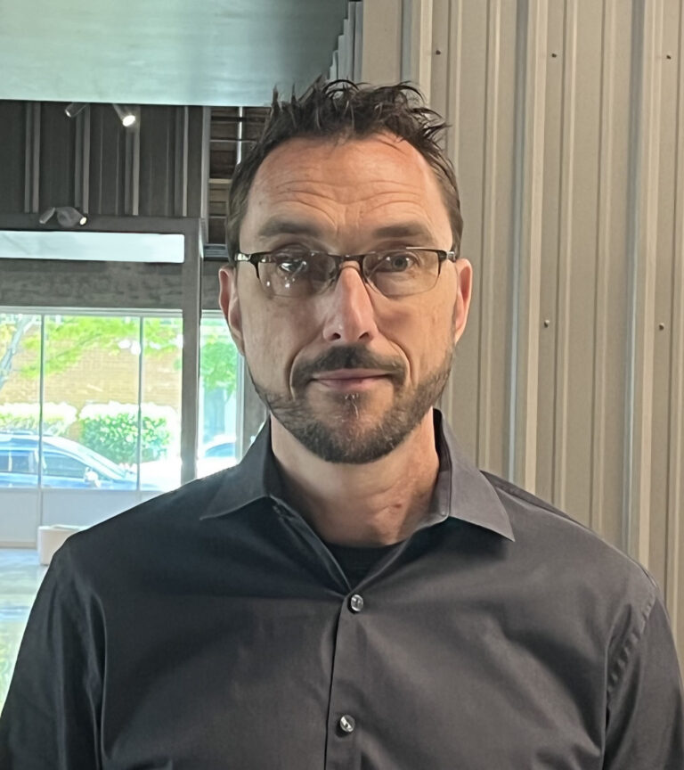 Man with glasses and a short beard in a dark button-up shirt, standing indoors near a window and paneled wall