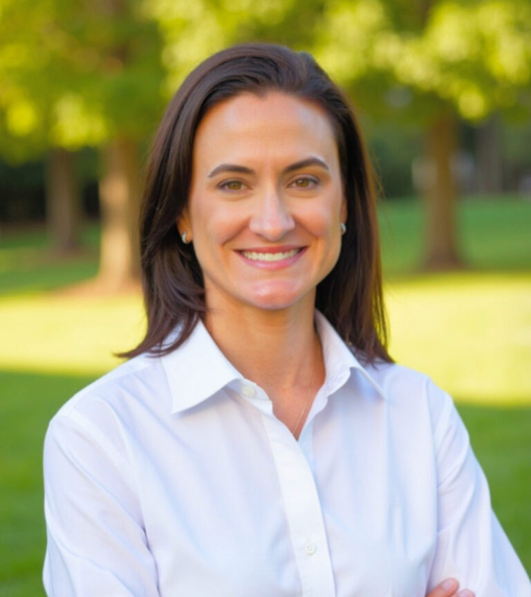 Smiling woman in a white button-down shirt standing outdoors with trees and grass in the background