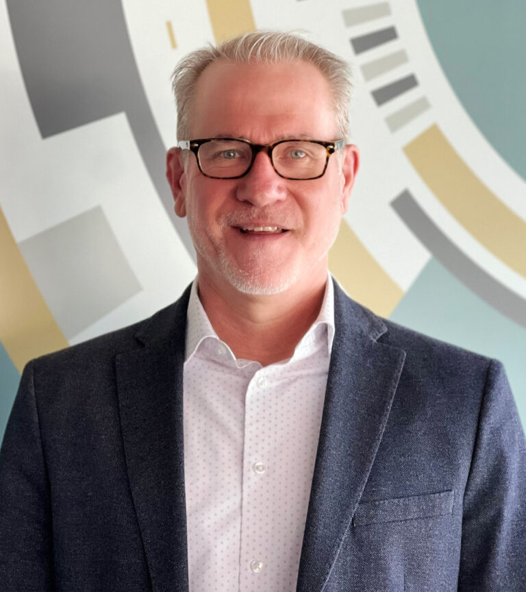 Smiling man in glasses wearing a dark blazer and white shirt, standing in front of a colorful geometric mural.